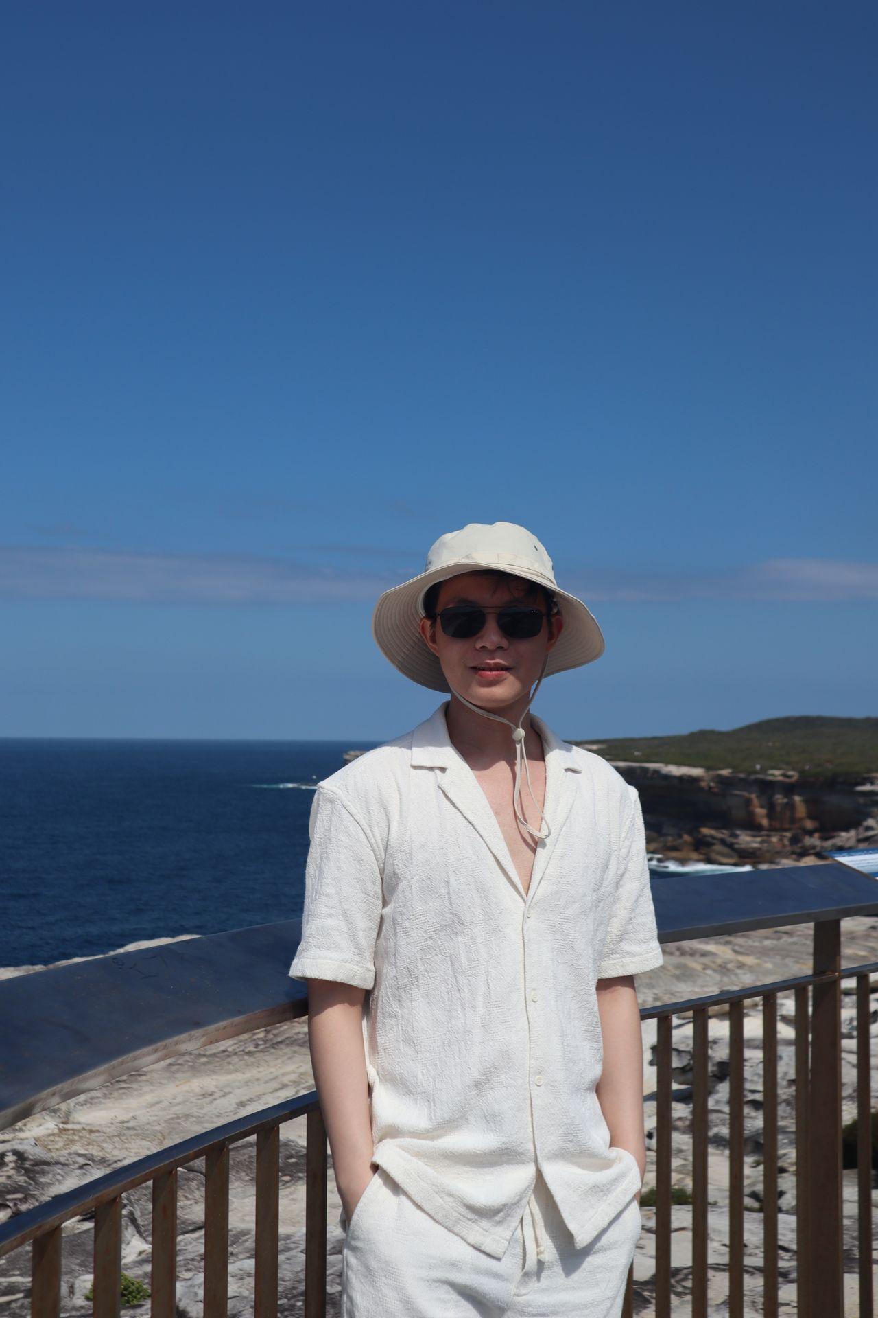 Person wearing a white outfit and hat standing near railing with ocean and cliffs in the background.